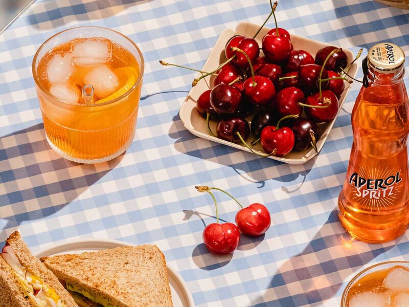 Centro Italia summer picnic scene featuring Aperol Spritz bottle, glass with ice and orange slice, fresh cherries, and sandwiches on a blue gingham tablecloth in sunlight, offering tips for the best Italian aperitivo experience.