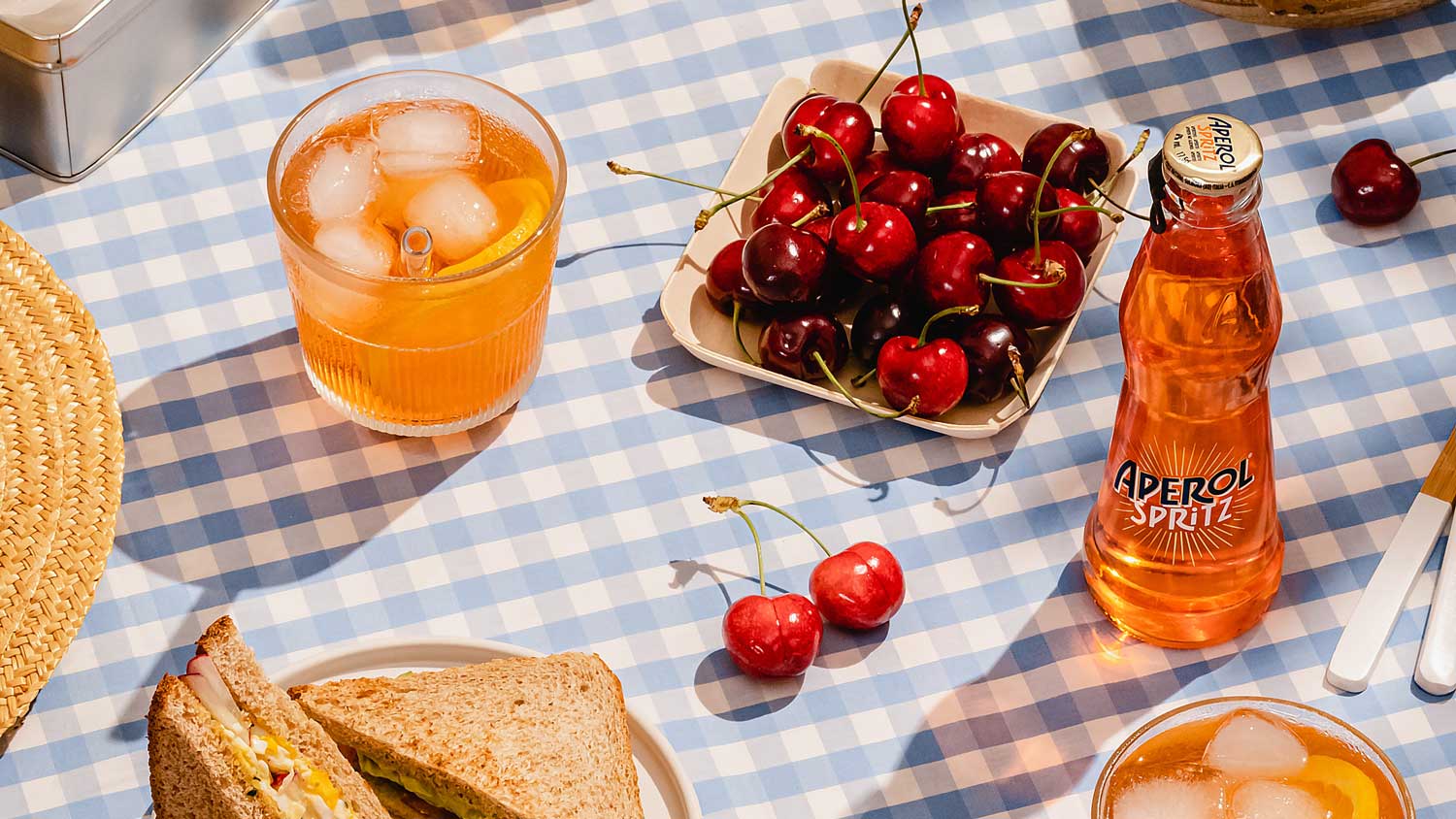 Centro Italia summer picnic scene featuring Aperol Spritz bottle, glass with ice and orange slice, fresh cherries, and sandwiches on a blue gingham tablecloth in sunlight, offering tips for the best Italian aperitivo experience.