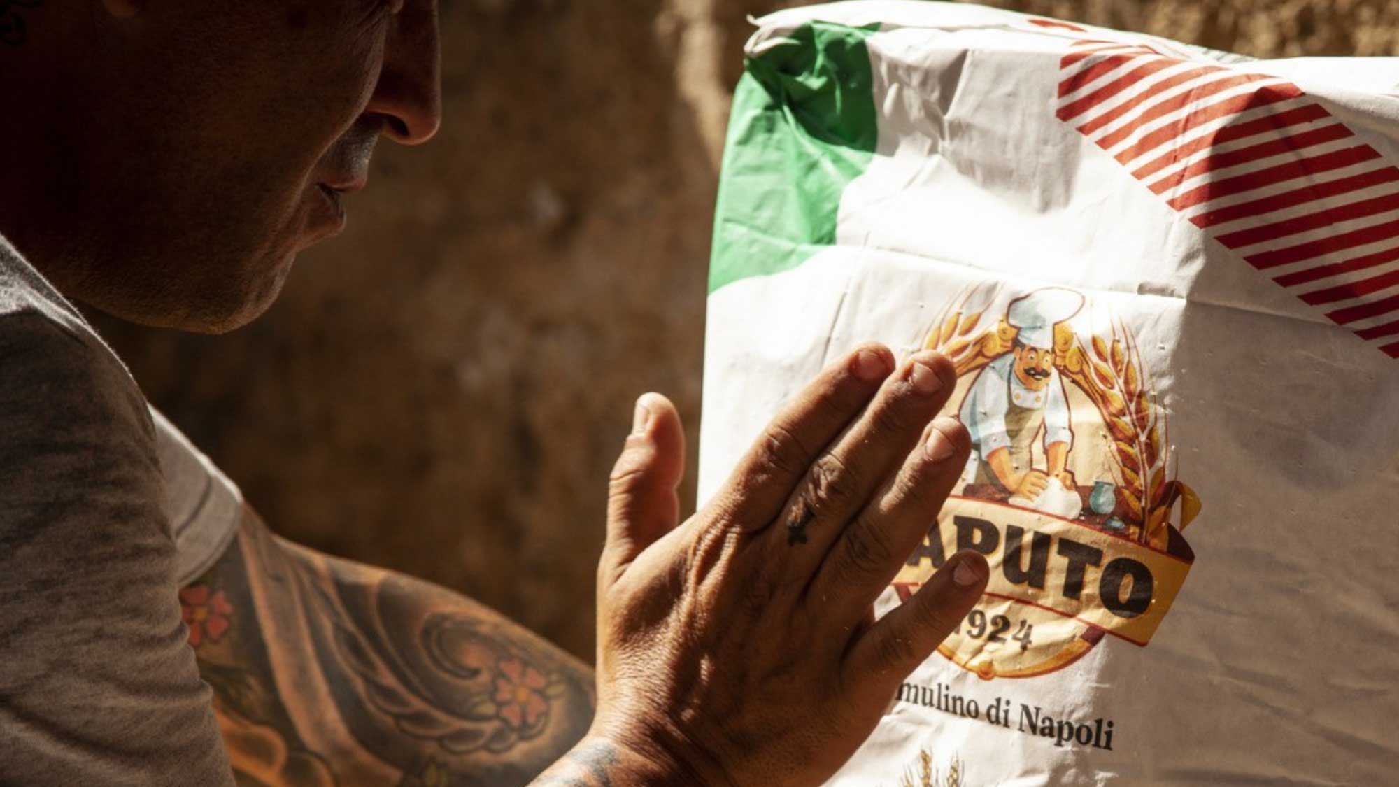 Ein tätowierter Mann legt seine Hand auf einen Mehlsack der Marke Caputo. Der Sack zeigt das bekannte Caputo-Logo, mit einem Bäcker und dem Schriftzug “Il mulino di Napoli - 1924”. Die Szene vermittelt eine enge Verbindung zur handwerklichen Tradition und zur hohen Qualität des Mehls, das in der italienischen Küche weit verbreitet ist