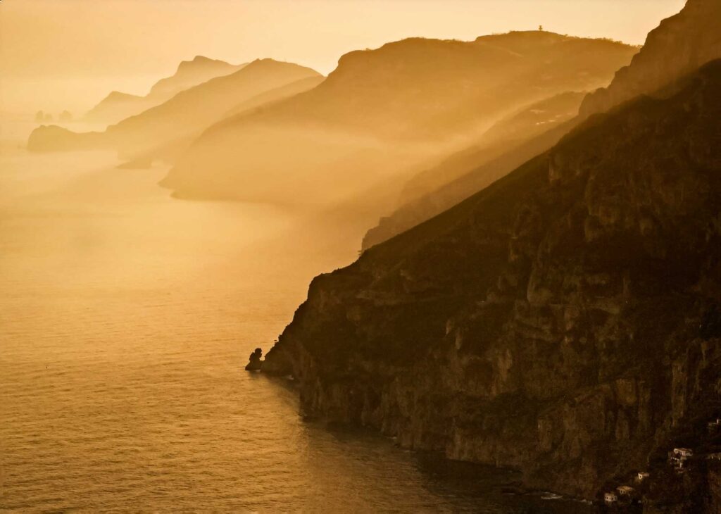 Eine beeindruckende, in Gold getauchte Küstenlandschaft Italiens, bei der die Hügel im Sonnenuntergang in den Nebel übergehen. Ein typisches Bild der Amalfiküste, bekannt für ihre Zitronenhaine und die Limoncello-Produktion