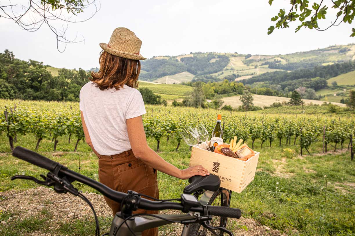 Eine Frau mit Strohhut schaut über die malerischen Weinberge des Weinguts Conte Vistarino, ein Picknickkorb mit Wein und Lebensmitteln ist auf ihrem Fahrrad platziert