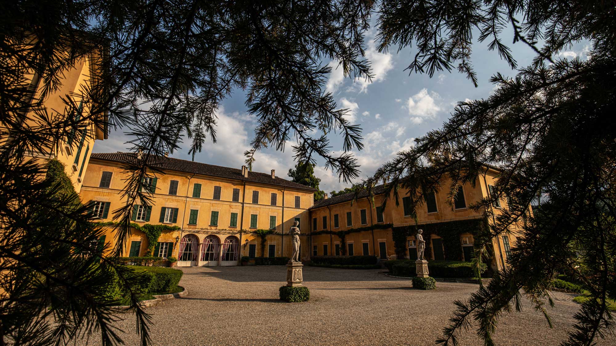 Der Innenhof des historischen Anwesens des Weinguts Conte Vistarino, eingerahmt von den Schatten der Bäume, mit gelben Gebäuden, grünen Fensterläden und klassischen Statuen, die den eleganten Charakter des Guts unterstreichen