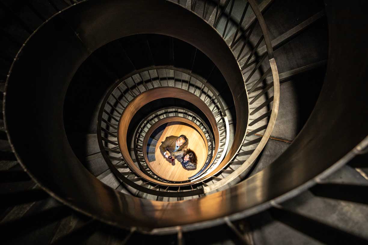 Ein Paar im spiralförmigen Treppenhaus des Weinguts Conte Vistarino, beide halten ein Weinglas und blicken nach oben