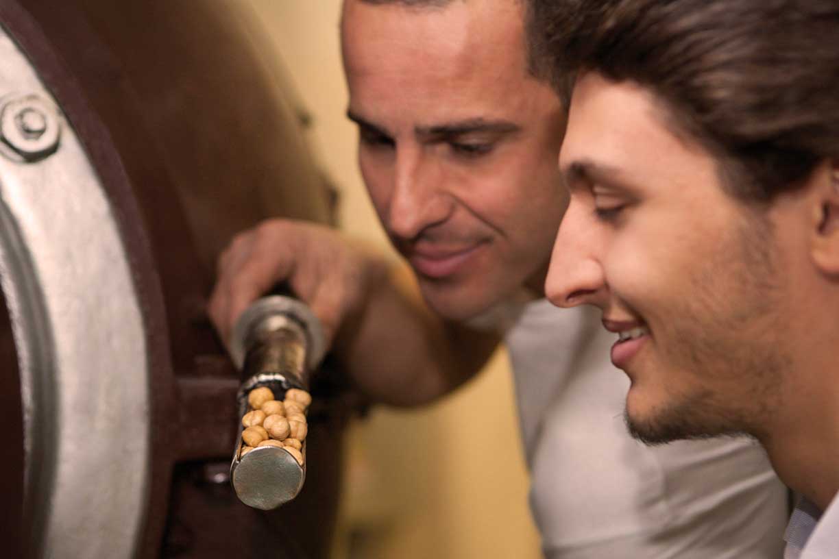 Zwei Handwerker, die Haselnüsse in einer Röstmaschine in der Fabrik von Antica Torroneria Piemontese genau inspizieren. Dies zeigt ihr Engagement, den Röstprozess für einen optimalen Geschmack in ihren Nougatprodukten zu perfektionieren