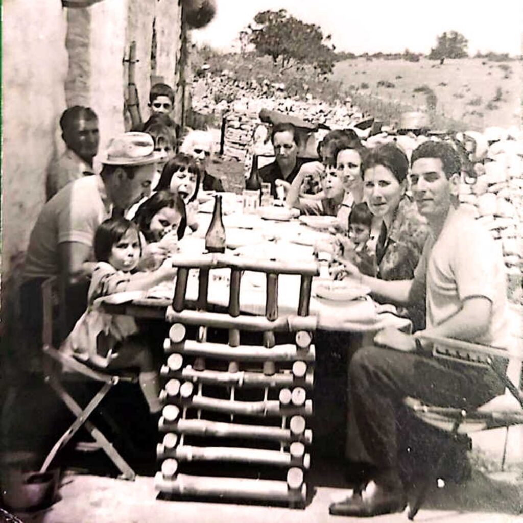 Ein historisches Schwarz-Weiß-Foto, das eine große italienische Familie bei einem gemeinsamen Essen im Freien zeigt. Die Szene spielt sich an einem langen Holztisch ab, der mit einfachen Speisen gedeckt ist. Im Hintergrund ist eine ländliche Umgebung mit Steinzäunen und Feldern zu sehen, die typisch für die Region Apulien ist, wo die berühmte Burrata-Käse-Tradition ihren Ursprung hat. Die entspannte und herzliche Atmosphäre unterstreicht die enge Verbindung zwischen Familie, Tradition und der handwerklichen Herstellung von Burrata.