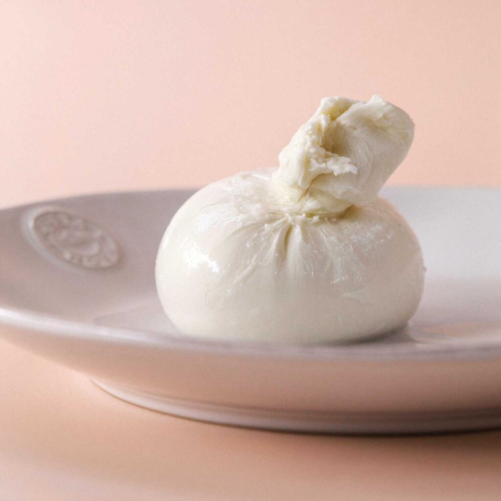 Eine minimalistische Nahaufnahme einer perfekt geformten Burrata auf einem eleganten weißen Teller, vor einem sanft rosafarbenen Hintergrund. Die glänzende Oberfläche des frischen italienischen Käseklassikers hebt die cremige Textur und die handgefertigte Qualität hervor. Der schlichte Hintergrund lenkt den Fokus auf die Burrata und betont ihre traditionelle Eleganz und ihren unverfälschten Geschmack.