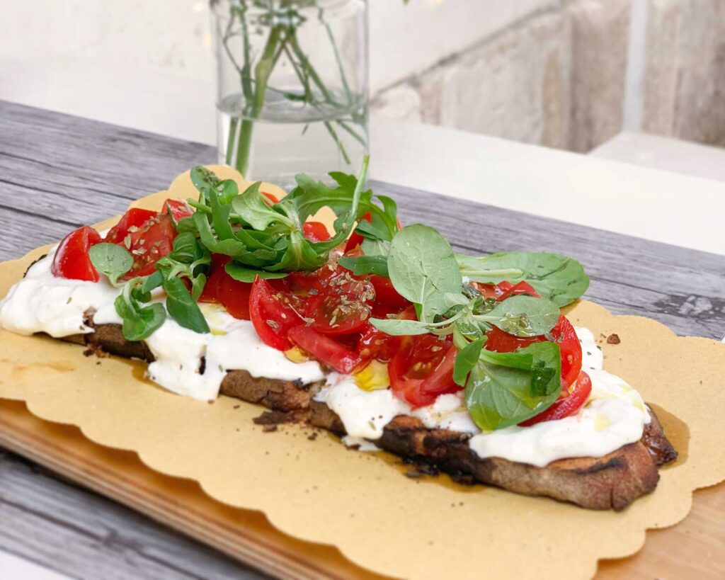 Eine köstlich angerichtete Burrata-Crostini auf rustikalem Holzbrett, garniert mit frischen Tomatenscheiben, Rucola und Basilikumblättern. Die cremige Burrata verschmilzt perfekt mit dem knusprigen Brot, während ein leichter Spritzer Olivenöl und Gewürze die Farben und Aromen hervorheben. Im Hintergrund steht eine Vase mit einer einzelnen Blume, die dem Setting eine entspannte mediterrane Atmosphäre verleiht.