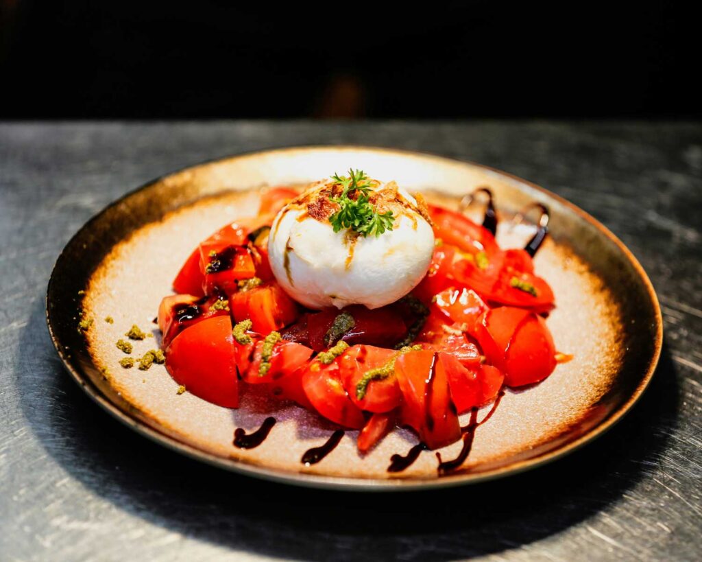 Eine kunstvoll angerichtete Burrata auf einem rustikalen Teller, umgeben von frischen, saftigen Tomatenstücken, verfeinert mit einem Hauch von grünem Pesto und Balsamico-Glasur. Die Burrata ist mit knusprigen Zwiebelstückchen und einem frischen Petersilienzweig dekoriert. Das Zusammenspiel von cremiger Burrata, süß-säuerlichem Balsamico und aromatischen Tomaten bietet einen harmonischen und ästhetischen Genussmoment.