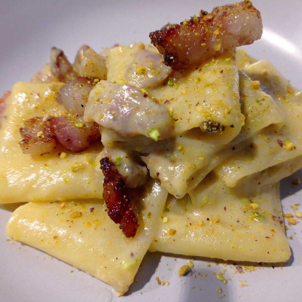 Eine Portion Paccheri-Nudeln mit Pistaziensauce, garniert mit gewürfeltem Pancetta und Pistazien, die die cremige Textur und die lebhaften Aromen in einer rustikalen italienischen Präsentation hervorheben.