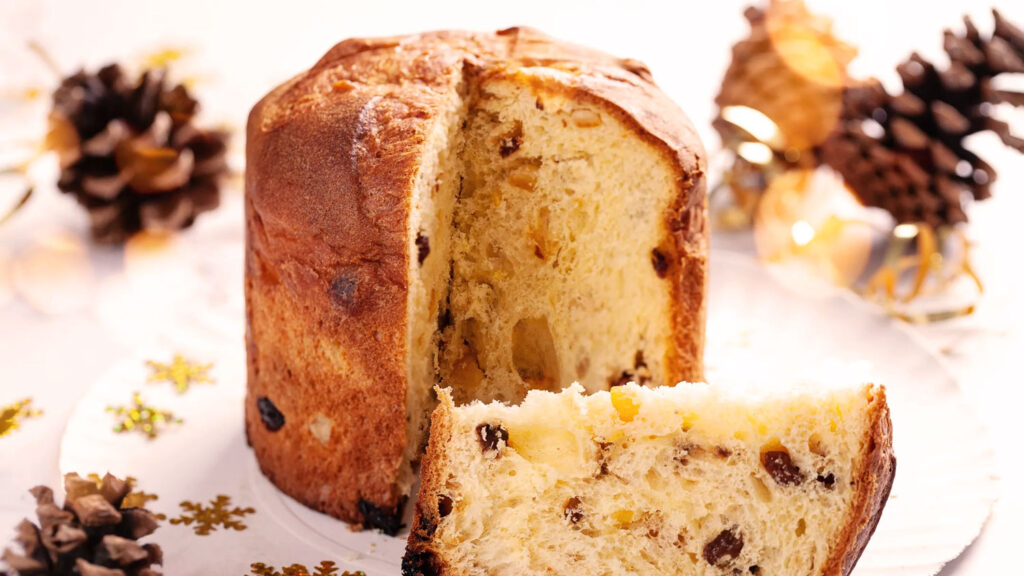 Ein goldbrauner, fluffiger Panettone mit einer weichen, luftigen Textur und reichlich eingebackenen Früchten und Nüssen ist angeschnitten, sodass das saftige Innere sichtbar wird. Der Panettone steht auf einem Teller, umgeben von weihnachtlicher Dekoration wie Tannenzapfen und glänzenden Ornamenten, die eine festliche Atmosphäre schaffen. Die Nahaufnahme unterstreicht die feine, traditionelle Zubereitung und die köstlichen Zutaten dieses italienischen Weihnachtsgebäcks