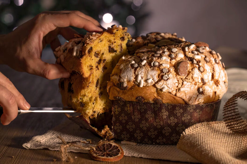 Eine Hand schneidet einen traditionellen Panettone an, dessen weiche, goldgelbe Krume mit Schokoladenstückchen durchzogen ist. Die oberste Kruste des Panettone ist mit Zucker, Mandeln und feinen Streuseln dekoriert, die ihm eine knusprige Textur verleihen. Der Panettone steht auf einem rustikalen Holzuntergrund, umgeben von natürlichen Dekorationen wie getrockneten Orangenscheiben und grobem Leinenstoff, was eine gemütliche und festliche Atmosphäre ausstrahlt