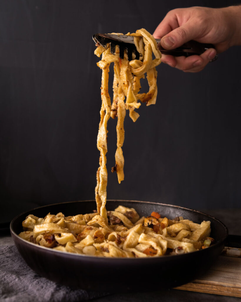 Frisch gebratene italienische Pasta in der Pfanne, mit einer Küchenzange hochgehoben – ein rustikales Gericht mit goldbraunen Nudeln und aromatischen Zutaten, perfekt für die authentische Küche Italiens.