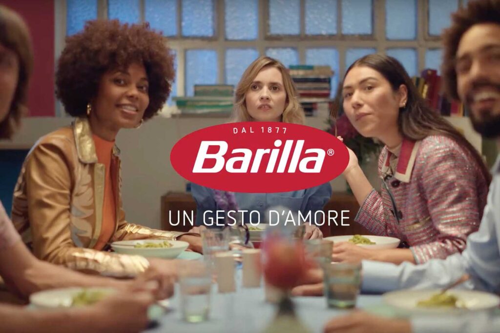 Eine Gruppe junger Menschen sitzt gemeinsam am Tisch und genießt eine Mahlzeit, die aus Pasta besteht. Im Vordergrund steht das Barilla-Logo mit dem Slogan „Un gesto d’amore“ (Ein Akt der Liebe). Die Szene vermittelt ein Gefühl von Zusammengehörigkeit und Freude am gemeinsamen Essen, unterstützt von Barillas Botschaft der Liebe und Wertschätzung, die durch das Teilen von Mahlzeiten zum Ausdruck kommt