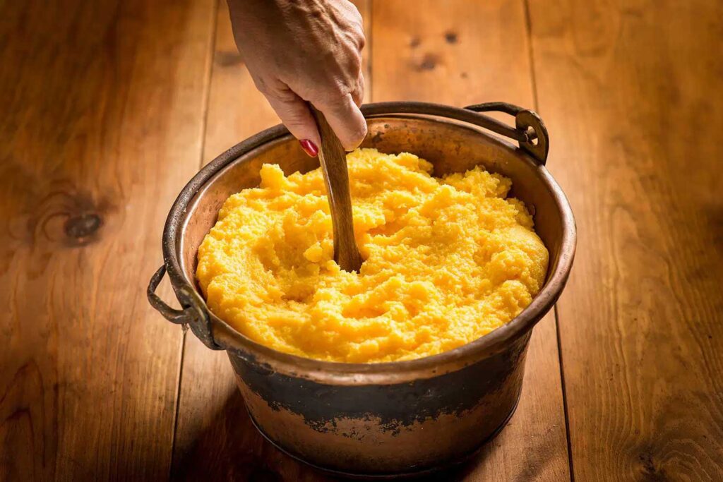 Ein rustikales Bild, das einen großen traditionellen Paiolo, einen Kupferkessel, zeigt, der mit frisch zubereiteter goldgelber Polenta gefüllt ist. Eine Hand mit rot lackierten Fingernägeln rührt die Polenta mit einem hölzernen Kochlöffel um. Der Paiolo steht auf einem Holztisch, dessen warme, natürliche Maserung zur gemütlichen Atmosphäre des Bildes beiträgt. Dieses Foto betont die handwerkliche Tradition und die Authentizität der Zubereitung von Polenta, einem zentralen Bestandteil der italienischen Küche.