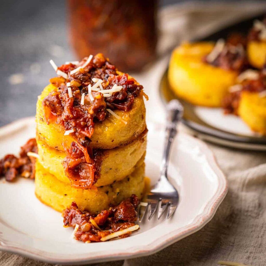 Ein kunstvoll angerichteter Teller mit drei perfekt gestapelten, goldbraun angebratenen Polentascheiben, die mit einer herzhaften Tomaten-Ragù-Sauce und geriebenem Käse garniert sind. Die Speise ist auf einem eleganten, weißen Porzellanteller angerichtet, daneben liegt eine silberne Gabel. Im Hintergrund verschwimmen weitere Portionen Polenta, die auf einem dunklen Teller serviert werden. Das Bild vermittelt eine moderne Interpretation von Polenta, die italienische Tradition mit raffinierter Präsentation verbindet.