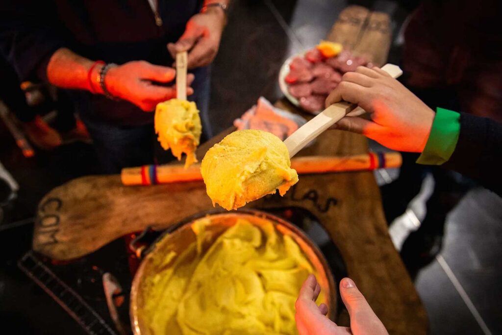 Eine lebendige Szene, in der mehrere Personen große Portionen frisch gekochter Polenta mit hölzernen Löffeln aus einem großen Topf schöpfen. Im Hintergrund sind ein rustikaler Holztisch und eine Platte mit verschiedenen Fleischsorten zu sehen, was die traditionelle italienische Geselligkeit und Kulinarik betont. Die warme Beleuchtung und die kräftigen Farben unterstreichen die Wärme und den Komfort dieses Moments, der die einfache, aber geschmackvolle Esskultur Italiens feiert.