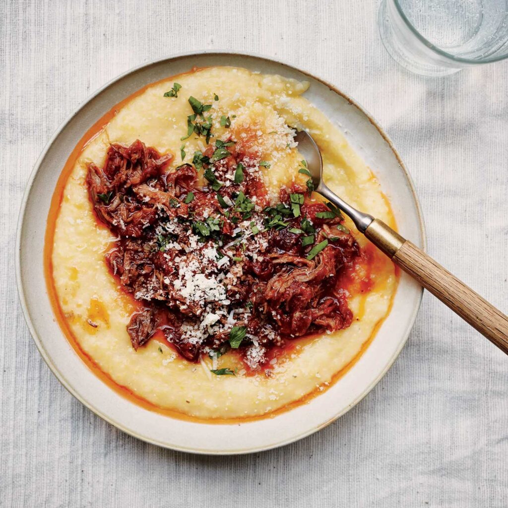 Ein Teller mit cremiger Polenta, gekrönt von einer herzhaften Tomatensauce mit zartem, geschmorten Fleisch, garniert mit frisch gehackter Petersilie und geriebenem Käse. Ein rustikaler Löffel mit Holzgriff liegt am Rand des Tellers. Das Gericht spiegelt die Einfachheit und den Reichtum der italienischen Küche wider, präsentiert auf einer hellen, strukturierten Tischdecke, die den Fokus auf das appetitliche Essen legt.