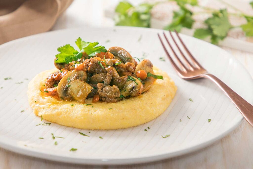 Ein elegantes Gericht aus cremiger Polenta, gekrönt mit einem herzhaften Ragout aus saftigen Pilzen, zarten Fleischstücken und knackigem Gemüse, garniert mit frischer Petersilie. Das Gericht ist kunstvoll auf einem weißen Teller angerichtet, neben einer glänzenden Kupfergabel, die einen Hauch von Raffinesse verleiht. Im Hintergrund sorgen unscharfe grüne Blätter und eine helle Tischdecke für eine warme und einladende Atmosphäre.