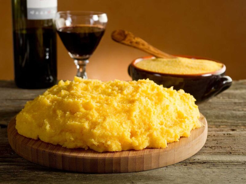 Ein rustikales Arrangement mit frisch zubereiteter Polenta, angerichtet auf einem runden Holzbrett. Im Hintergrund steht eine traditionelle schwarze Schale mit Polenta und einem Holzlöffel, sowie ein Glas tiefroten Weins und eine Weinflasche, die das italienische Flair unterstreichen. Die Szene ist auf einem rustikalen Holztisch präsentiert, mit warmem Licht, das die leuchtend gelbe Farbe der Polenta hervorhebt. Dieses Bild vermittelt die Authentizität und den Genuss italienischer Tradition.