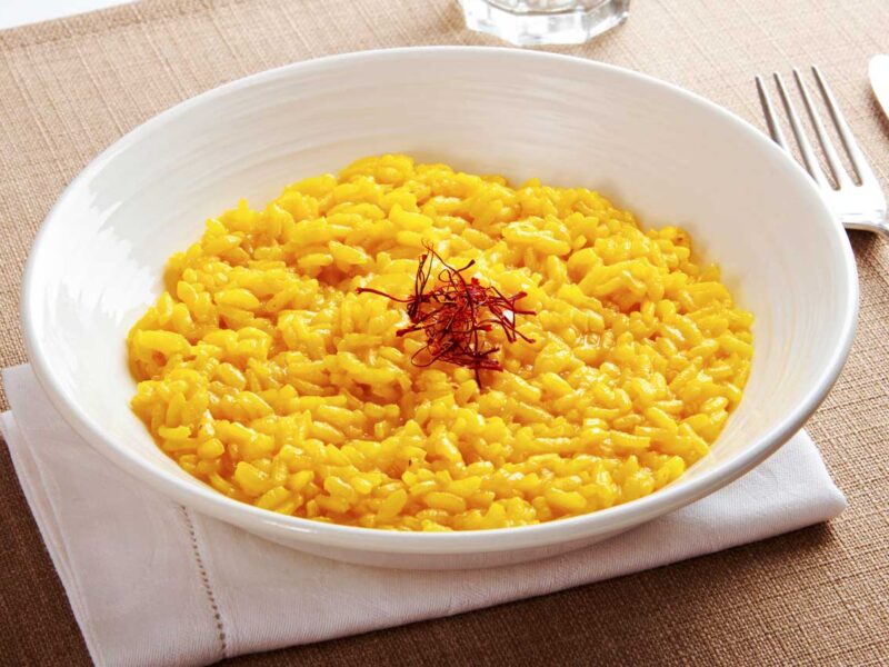 Risotto Milanese mit Safran. Ein Teller mit leuchtend gelbem Risotto Milanese, verfeinert mit Safranfäden in der Mitte, präsentiert auf einer weißen Serviette. Dieses traditionelle italienische Gericht wird auf der Centro Italia Gazzetta als Rezept vorgestellt