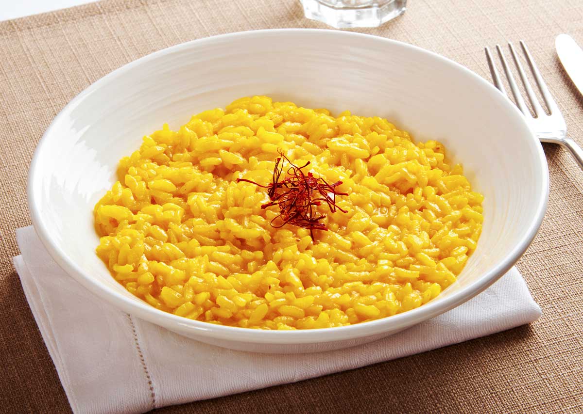 Risotto Milanese mit Safran. Ein Teller mit leuchtend gelbem Risotto Milanese, verfeinert mit Safranfäden in der Mitte, präsentiert auf einer weißen Serviette. Dieses traditionelle italienische Gericht wird auf der Centro Italia Gazzetta als Rezept vorgestellt