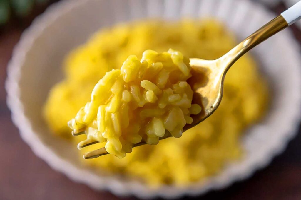 Nahaufnahme eines goldenen Löffels, der eine Portion cremigen Risotto Milanese mit Safranfärbung aufnimmt. Im Hintergrund ist ein Teller mit dem leuchtend gelben Risotto zu sehen. Diese Abbildung illustriert ein Kochrezept für “Risotto milanese allo Zafferano” auf der Centro Italia Gazzetta