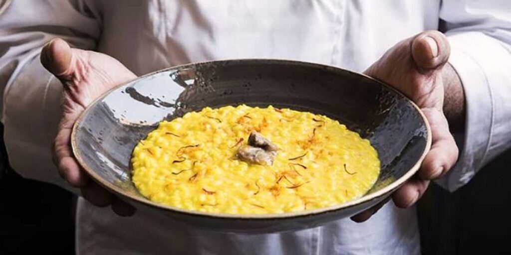 Ein Teller mit cremigem, gelbem Risotto Milanese mit Safran, angerichtet mit einem kleinen Stück Mark in der Mitte, wird von einem Koch in weißen Ärmel gehalten. Die Darstellung begleitet ein Rezept für “Risotto milanese allo Zafferano” auf der Centro Italia Gazzetta und vermittelt die warme, traditionelle Atmosphäre dieses italienischen Klassikers