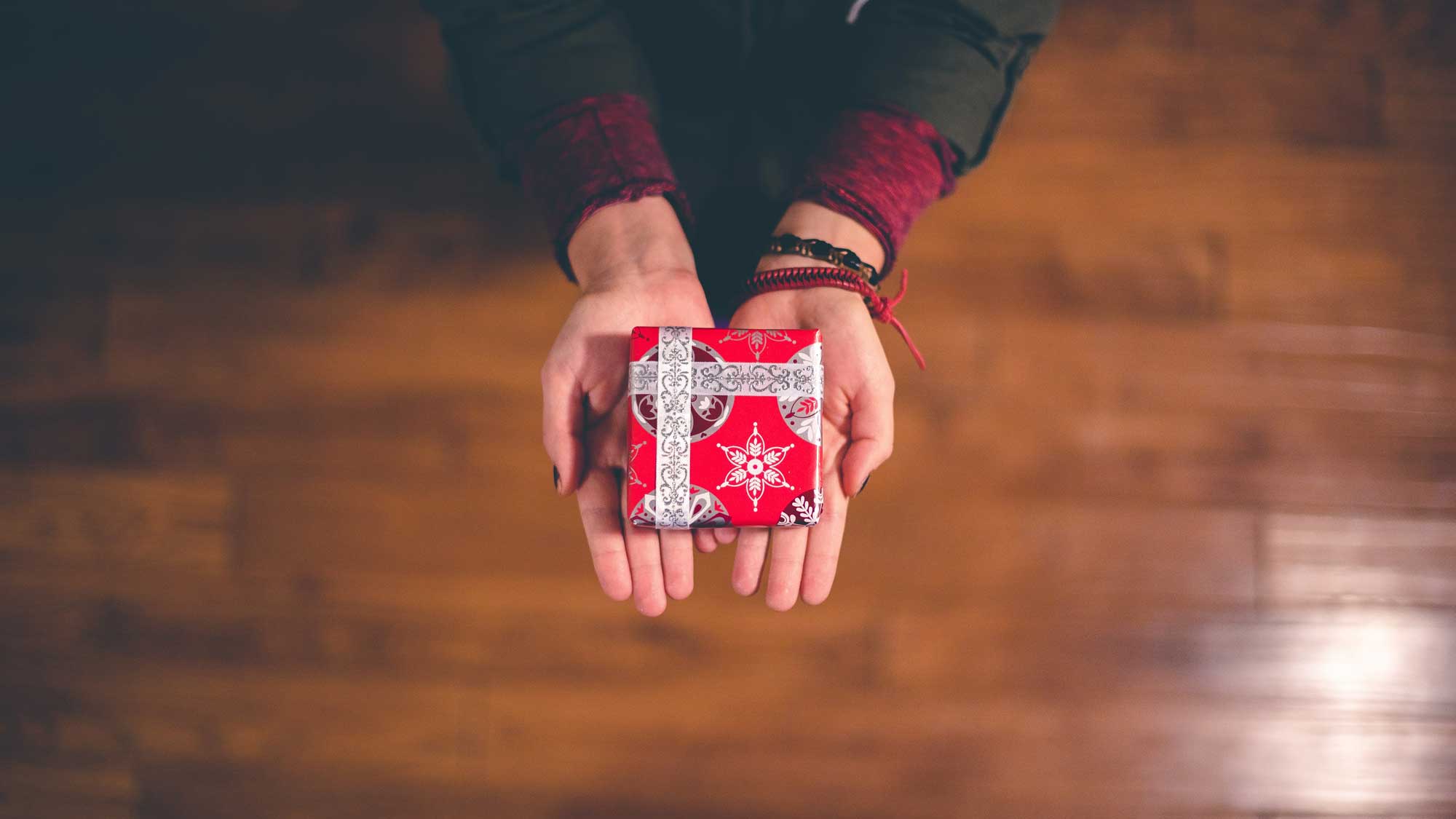 Zwei Hände halten ein kleines, festlich verpacktes Geschenk mit rotem Papier und dekorativen Mustern, perfekt als Symbol für Gutscheine und Geschenke, wie sie auf der Seite “Centro Italia Gutscheine” angeboten werden.