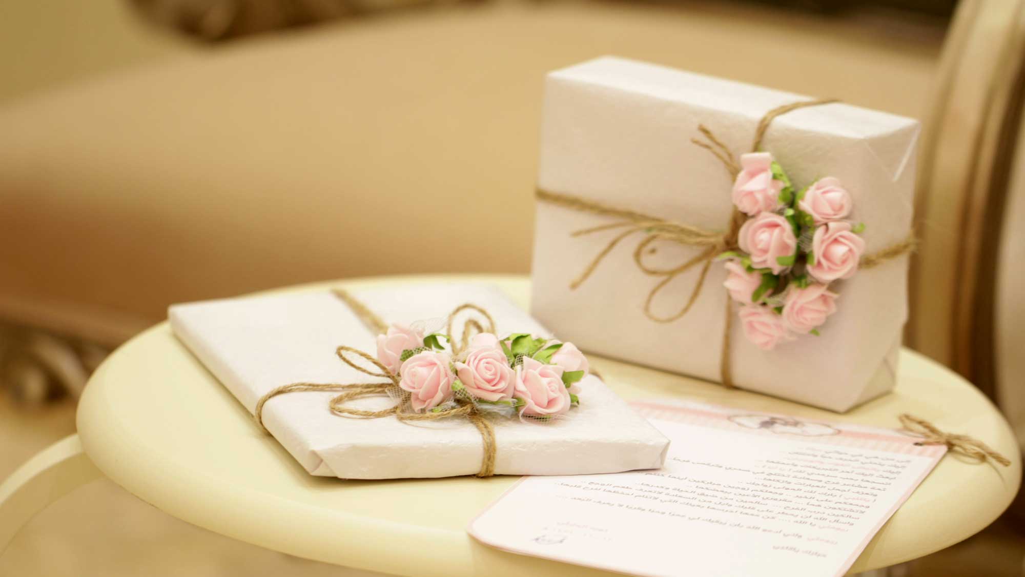 Zwei elegant verpackte Geschenke mit weißem Papier, dekoriert mit rosa Blumen und Juteband, stehen auf einem cremefarbenen Tisch. Ein Brief liegt daneben, symbolisch für die Geschenkideen und Gutscheine von Centro Italia.