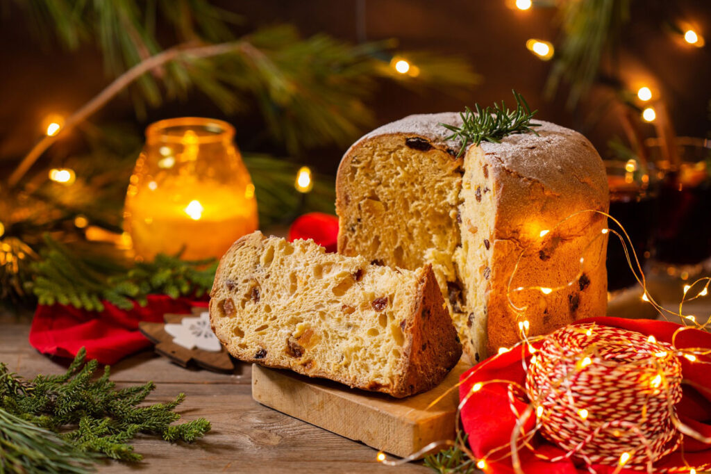 Ein aufgeschnittener Panettone, umgeben von weihnachtlicher Dekoration, darunter Tannenzweige, Kerzenlicht und ein rotes Band, das für festliche Atmosphäre sorgt.