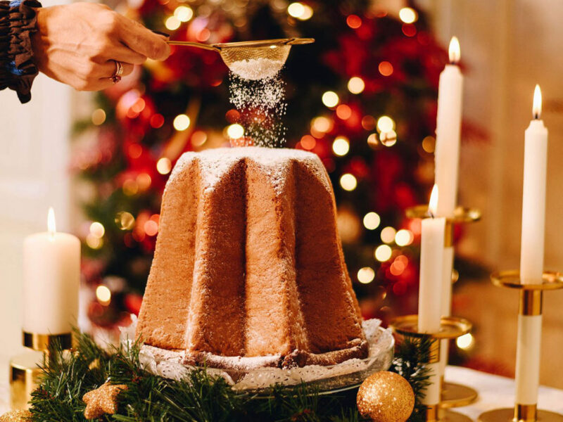 Ein Pandoro, der mit Puderzucker bestreut wird, umgeben von Weihnachtsdekoration wie Kerzen, Tannenzweigen und einem geschmückten Weihnachtsbaum im Hintergrund.