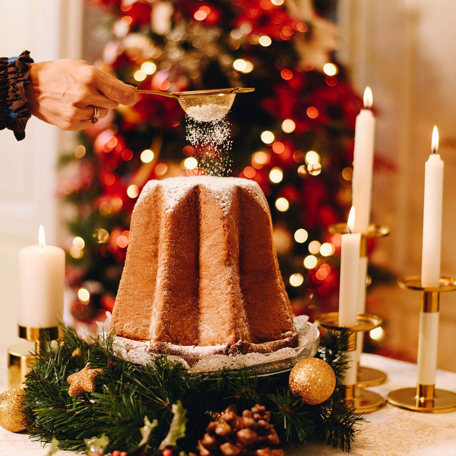 Ein Pandoro, der mit Puderzucker bestreut wird, umgeben von Weihnachtsdekoration wie Kerzen, Tannenzweigen und einem geschmückten Weihnachtsbaum im Hintergrund.