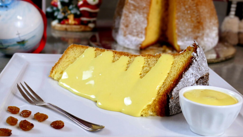 Ein Stück saftiger Pandoro, großzügig mit einer cremigen Zabaione-Sauce überzogen, auf einem weißen Teller serviert. Im Hintergrund weihnachtliche Dekorationen mit einem festlichen Flair.