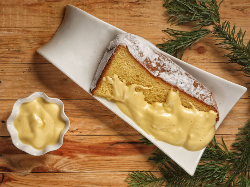 Eine festliche Servierplatte mit einem großen Stück Pandoro, bedeckt mit cremiger Zabaione-Sauce. Daneben ein kleines Schälchen mit Zabaione und Tannenzweigen, die eine weihnachtliche Atmosphäre schaffen.