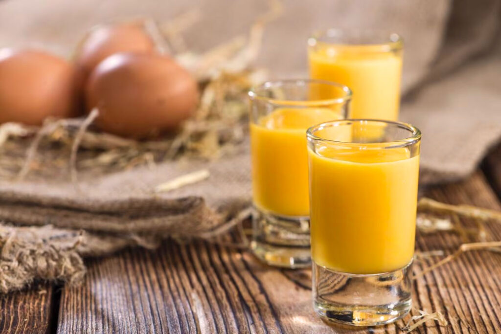 Drei kleine Gläser mit Eierlikör stehen auf einem rustikalen Holztisch, begleitet von braunen Eiern im Hintergrund – eine Hommage an die traditionelle Herstellung von Bombardino.