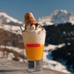 Ein Bombardino-Drink, garniert mit Sahne und einem Schuss Kakao, präsentiert vor der atemberaubenden Kulisse der verschneiten italienischen Alpen – das perfekte Après-Ski-Getränk.