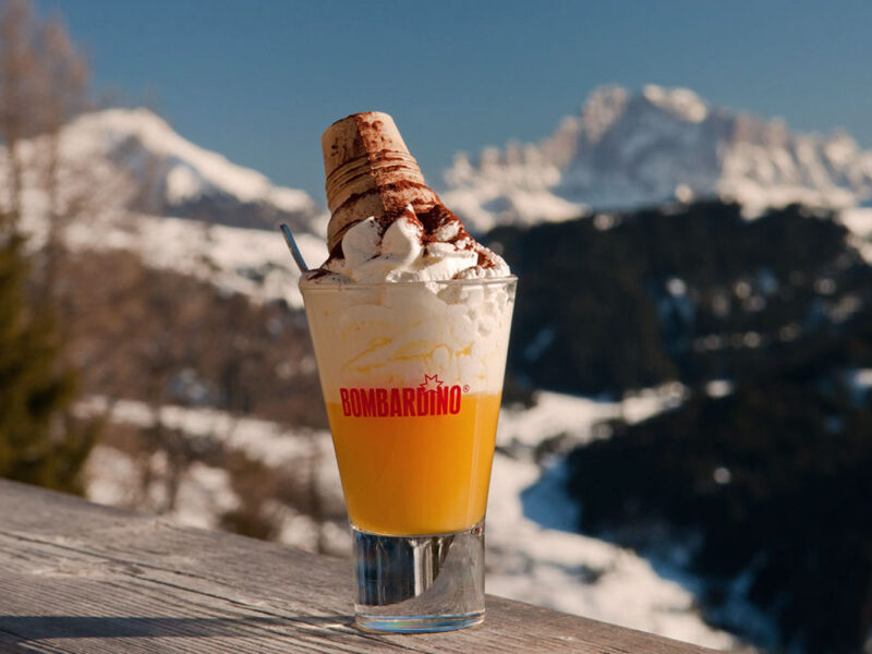 Ein Bombardino-Drink, garniert mit Sahne und einem Schuss Kakao, präsentiert vor der atemberaubenden Kulisse der verschneiten italienischen Alpen – das perfekte Après-Ski-Getränk.