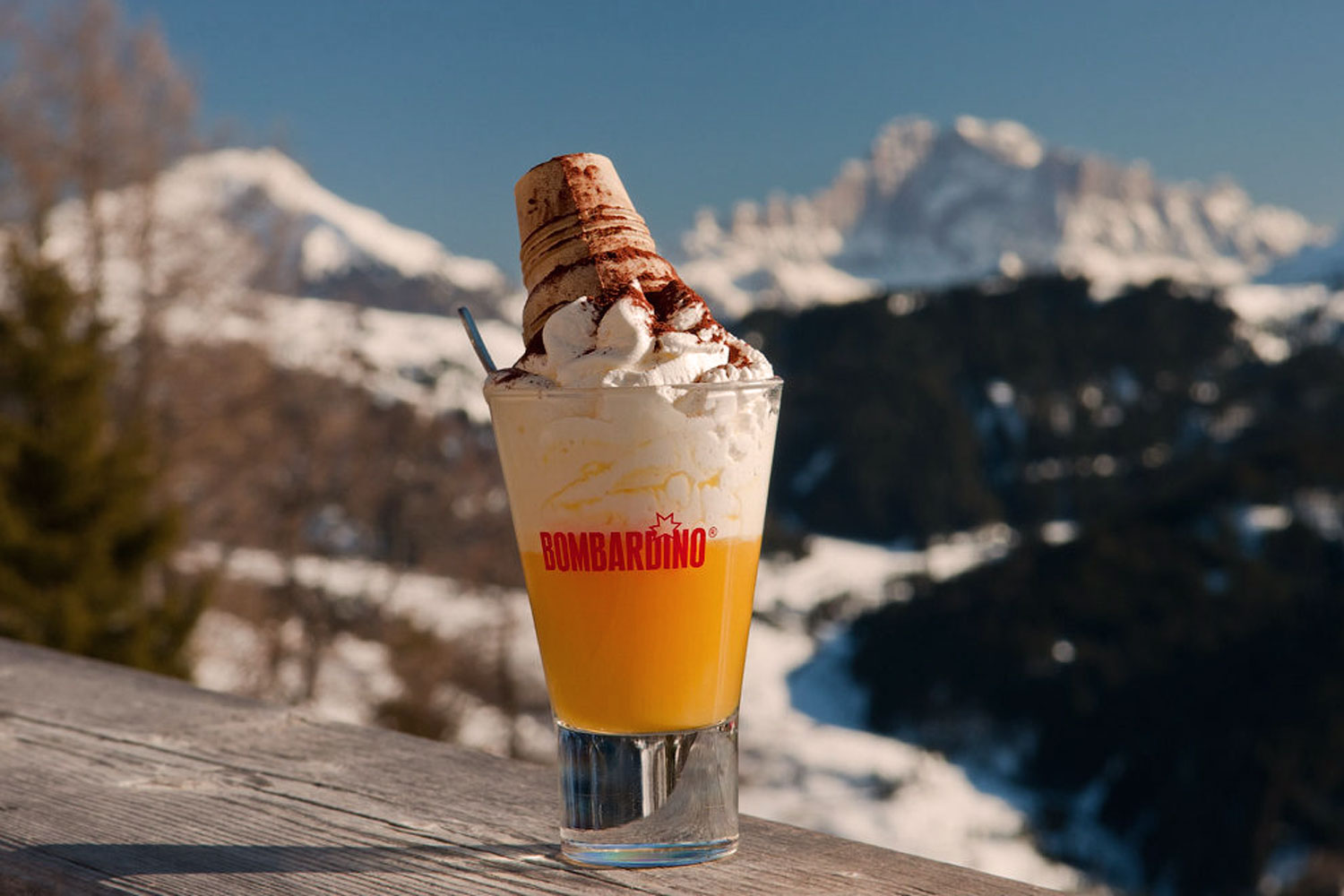 Ein Bombardino-Drink, garniert mit Sahne und einem Schuss Kakao, präsentiert vor der atemberaubenden Kulisse der verschneiten italienischen Alpen – das perfekte Après-Ski-Getränk.