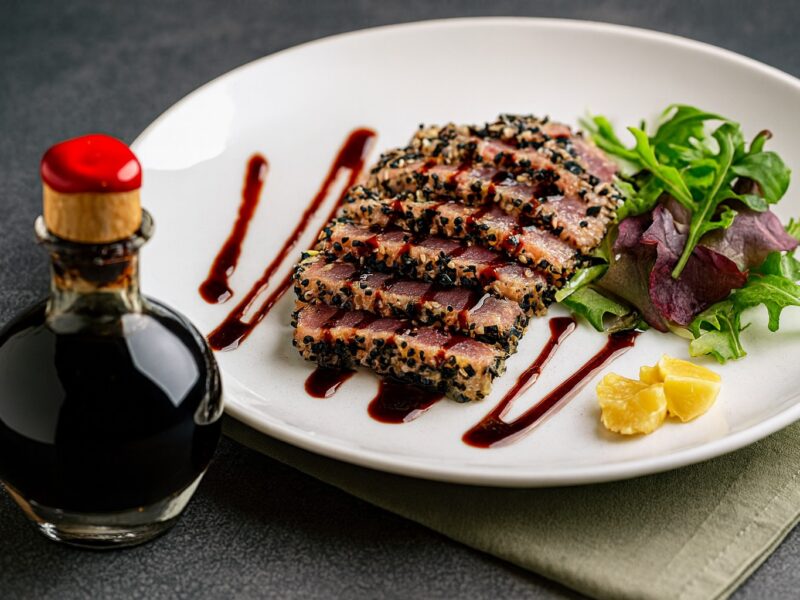 Scheiben von Sesamkrusten-Thunfisch mit frischem Blattsalat und Balsamico-Reduktion auf weißem Teller, daneben eine dekorative Glasflasche Aceto Balsamico.