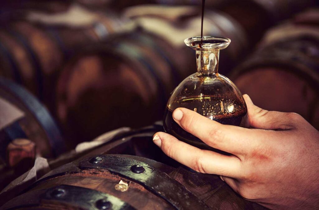 Ein Hand hält eine Glasflasche mit dunklem Aceto Balsamico, während der Essig in die Flasche tropft. Im Hintergrund mehrere Holzfässer in einer traditionellen Essigmanufaktur.