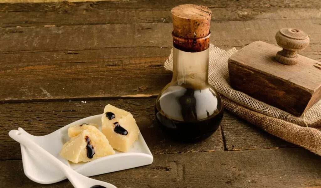 Eine Flasche traditioneller Aceto Balsamico auf einem rustikalen Holztisch. Daneben liegen kleine Stücke Parmesankäse mit einem Hauch von dunklem Balsamico-Essig verziert. Ein Holzsiegel und grobes Leinenstoff ergänzen die authentische Darstellung.