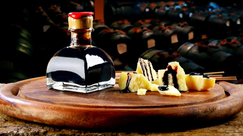 Edler Aceto Balsamico mit Parmigiano Reggiano: Eine elegante Glasflasche mit dunklem, gereiftem Balsamico-Essig steht auf einem rustikalen Holzbrett. Daneben liegen Stücke von Parmigiano Reggiano, kunstvoll mit Balsamico beträufelt. Im Hintergrund sind traditionelle Holzfässer zur Essigreifung sichtbar.