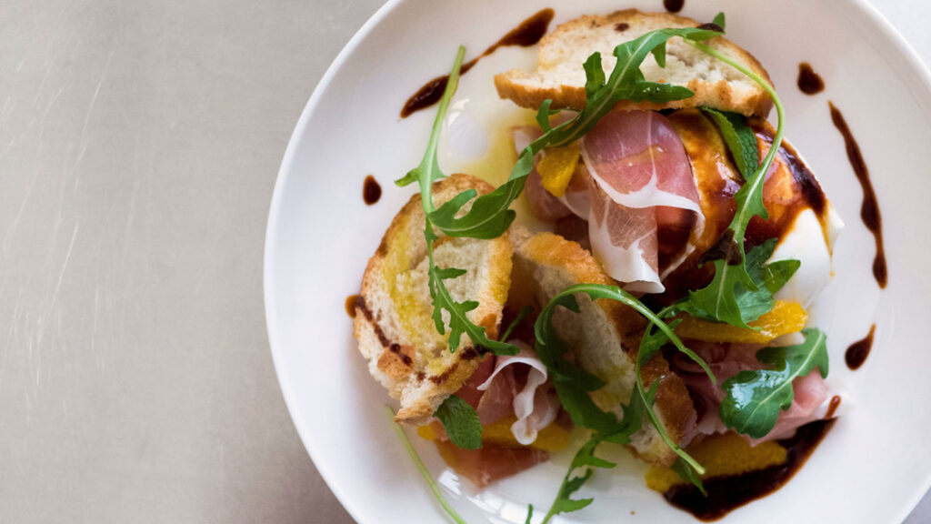 Ein Gourmet-Teller mit knusprigem Brot, Prosciutto, Mozzarella, Rucola und Orangenstücken, verfeinert mit Aceto Balsamico – eine raffinierte Kombination aus süßen, salzigen und sauren Aromen.