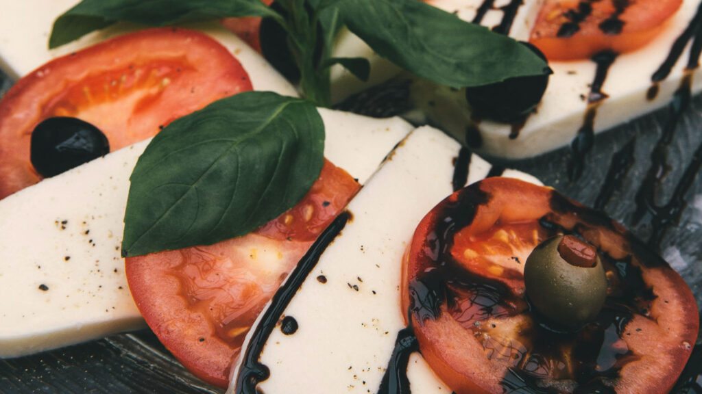 Nahaufnahme eines klassischen Caprese-Salats mit Tomaten, Mozzarella und frischem Basilikum, verfeinert mit Aceto Balsamico und schwarzer Olive.