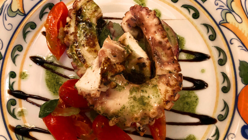 Mediterrane Vorspeise mit gegrilltem Oktopus, Kirschtomaten und Basilikum-Pesto, kunstvoll mit Aceto Balsamico garniert.