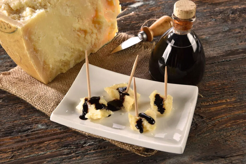 Stücke von gereiftem Parmigiano Reggiano mit Aceto Balsamico beträufelt, serviert auf einem weißen Teller mit Zahnstocher-Spießen. Im Hintergrund eine Flasche des edlen Essigs und ein Käsemesser auf rustikalem Holztisch.