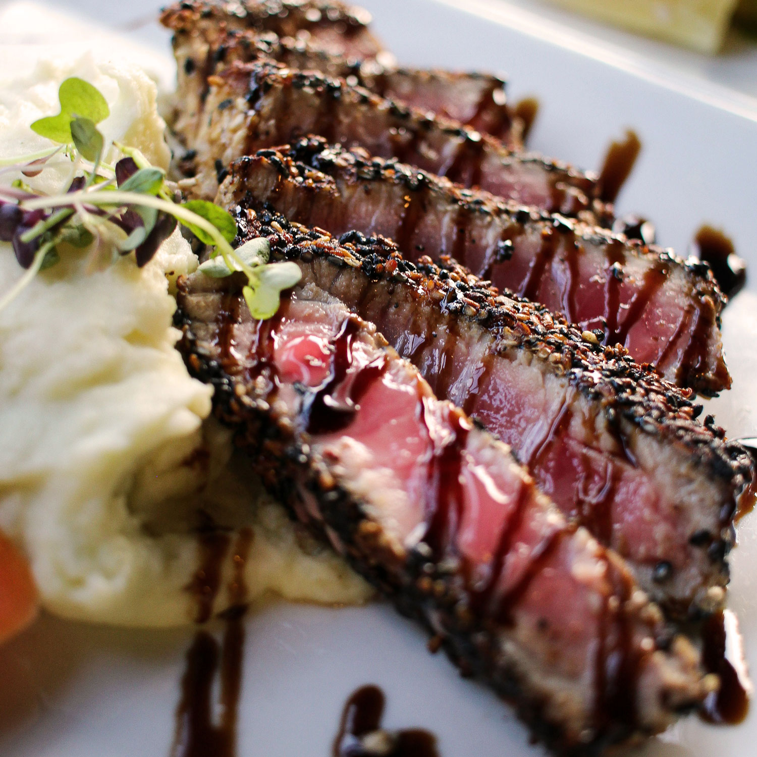 Ein Teller mit in Sesamkruste gebratenem, rosa-gegartem Thunfischfilet, serviert mit Kartoffelpüree und mit Aceto Balsamico beträufelt.