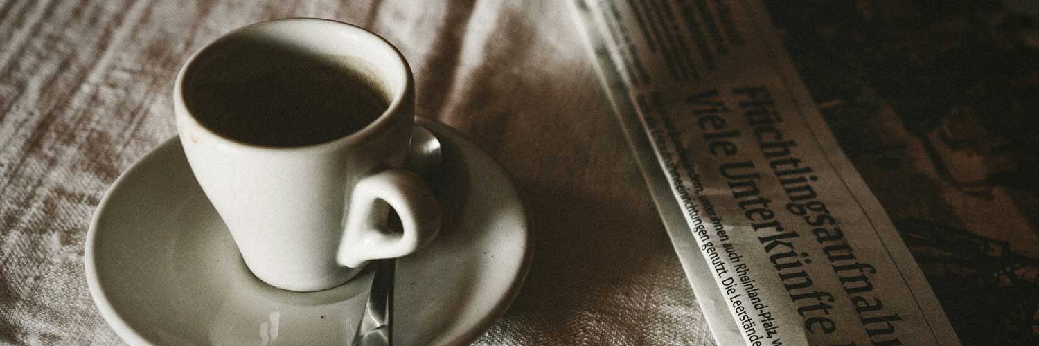 Eine Tasse Espresso auf einem Unterteller mit Löffel, daneben eine aufgeschlagene Zeitung mit sichtbarer Schlagzeile. Stimmungsvolle, nostalgische Atmosphäre – Symbolbild für die Presse- und Nachrichtenrubrik von Centro Italia