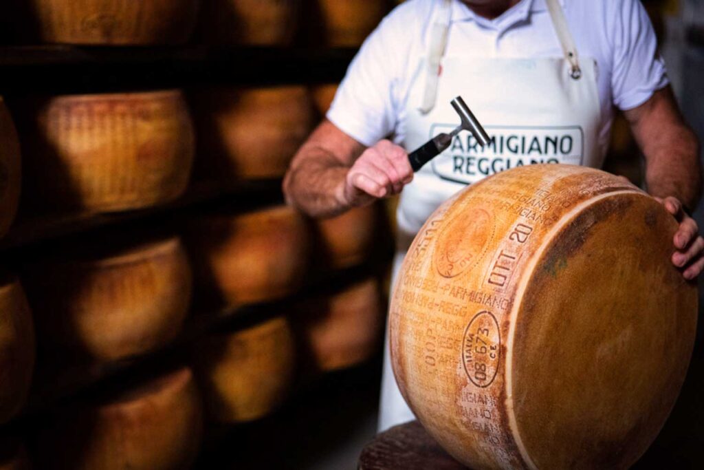 Ein Käsemeister prüft einen Laib Parmigiano Reggiano mit einem speziellen Metallhammer, um die Qualität und Reife des Käses zu bestimmen. Im Hintergrund sind weitere gereifte Laibe in einem Käselager zu sehen.