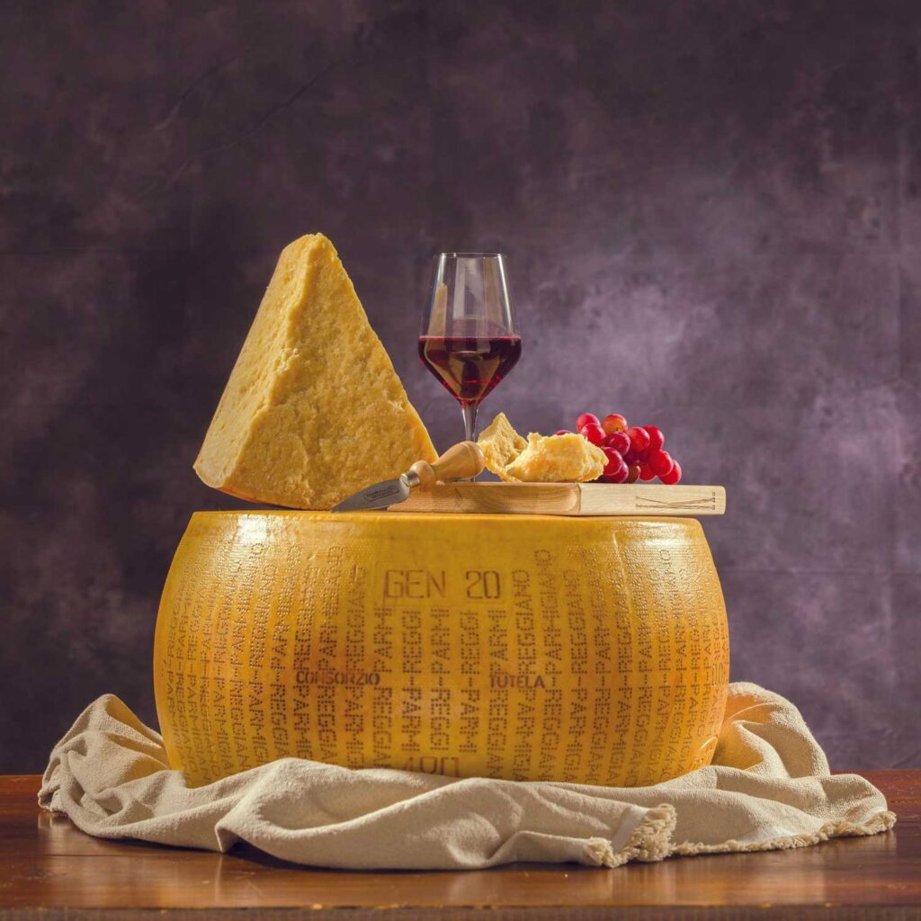 Ein großes Rad Parmigiano Reggiano mit einem Dreieckstück darauf, begleitet von einem Glas Rotwein, Trauben und Brot auf einem Holzbrett. Der Käse liegt auf einem Leinentuch vor einem dunklen, strukturierten Hintergrund – eine perfekte Darstellung italienischer Genusskultur.